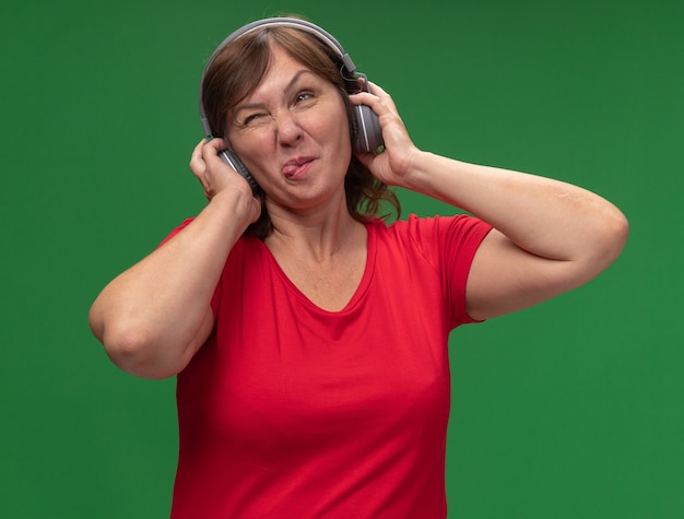 Kobieta w średnim wieku w czerwonej koszulce ze słuchawkami wystający język szczęśliwa i radosna stojąca nad zieloną ścianą