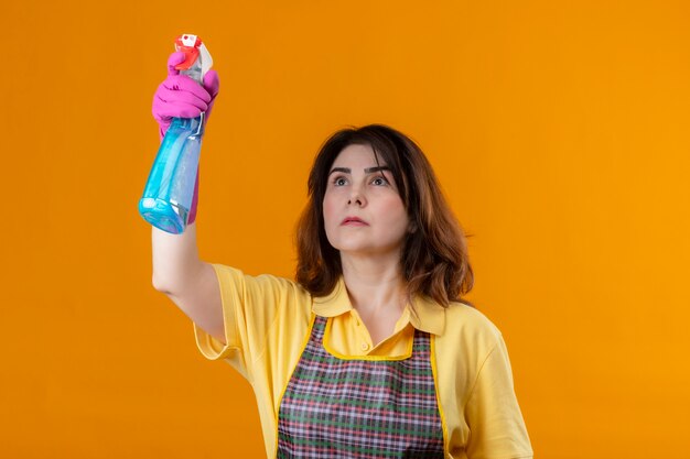 Kobieta w średnim wieku w fartuchu i gumowych rękawiczkach do czyszczenia za pomocą sprayu do czyszczenia z poważną twarzą stojącą na pomarańczowej ścianie