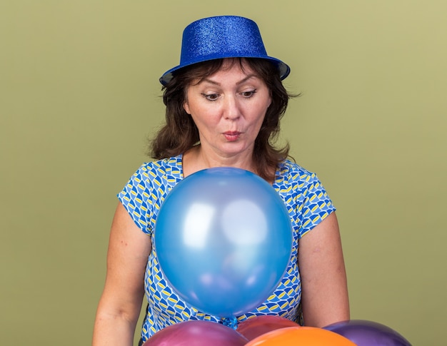 Kobieta w średnim wieku w imprezowym kapeluszu z wiązką kolorowych balonów wygląda na zdziwioną i zaskoczoną świętującą przyjęcie urodzinowe stojące nad zieloną ścianą