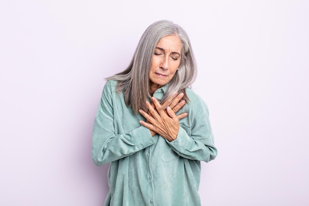 Kobieta w średnim wieku z siwymi włosami, smutna, zraniona i załamana, trzymająca obie ręce blisko serca, płacząca i przygnębiona