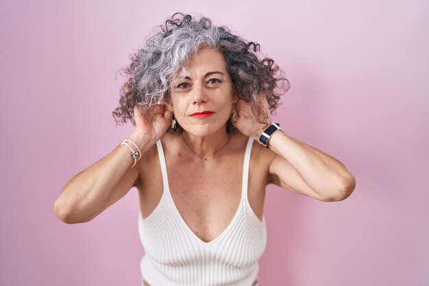 Kobieta w średnim wieku z siwymi włosami stojąca na różowym tle, próbująca usłyszeć gest obu rąk na uchu, ciekawa plotek. problem ze słuchem, głuchy
