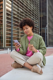 Kobieta w stroju codziennym siada krzyżuje nogi używa telefonu komórkowego komunikuje się w internecie posty informacje w mediach społecznościowych napoje aromatyczna kawa z jednorazowego kubka pozuje na zewnątrz