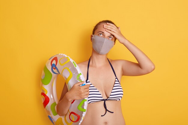Kobieta w stroju kąpielowym trzymająca w rękach gumowy pierścień, ubrana w higieniczną maskę zapobiegającą zakaźnemu wirusowi na plaży kurortu, kobieta wyglądająca na zmęczoną, trzymająca rękę na czole.