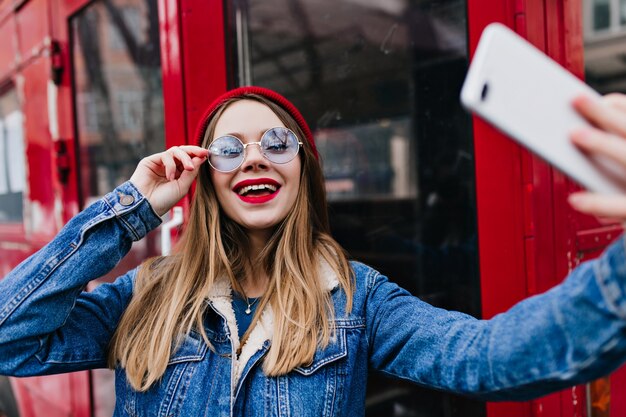 kobieta w stylowej kurtce dżinsowej przy użyciu telefonu do selfie i śmiejąc się.