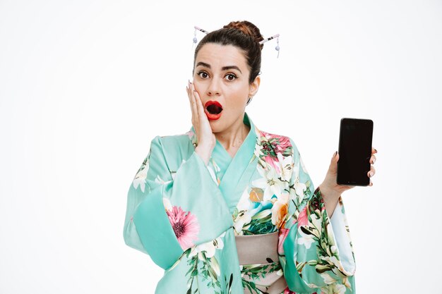 Kobieta w tradycyjnym japońskim kimonie pokazująca smartfona zdumiona i zdziwiona na białym tle