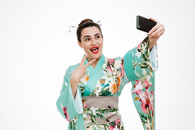 Kobieta w tradycyjnym japońskim kimonie szczęśliwa i pozytywnie uśmiechnięta pokazująca znak v robi selfie za pomocą smartfona na białym