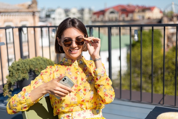 Kobieta w żółtej sukience na tarasie w letniej kawiarni z telefonem komórkowym w słoneczny dzień, wygląda szczęśliwie i pozytywnie z ogromnym uśmiechem na twarzy