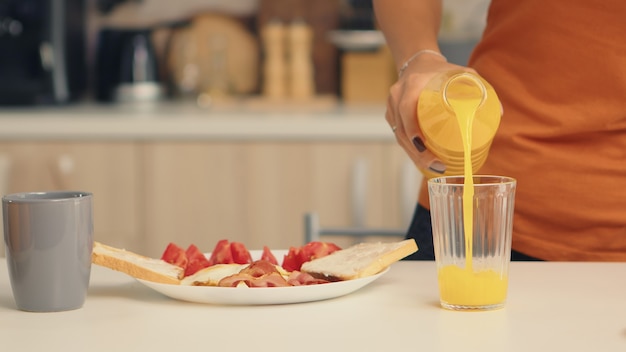 Kobieta wlewając świeży sok do szklanki na śniadanie w kuchni. Kobieta pije zdrowy i naturalny sok pomarańczowy. Gospodyni pijąca zdrowy, naturalny, domowy sok pomarańczowy. Orzeźwiający niedzielny poranek