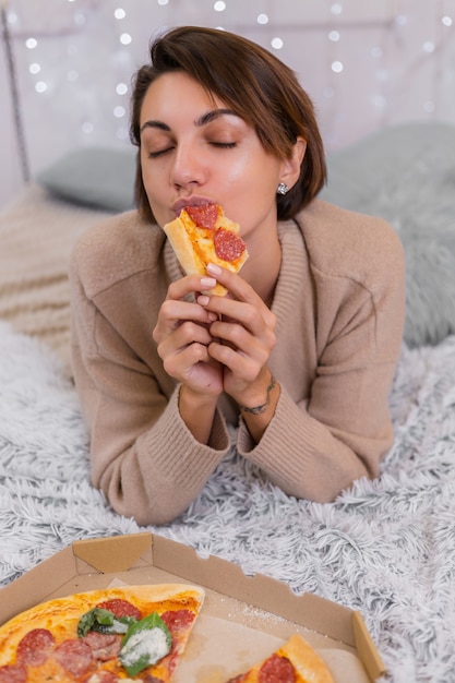 Kobieta wschód fast food od dostawy na łóżku w sypialni w domu. Kobieta sama ciesząc się tłustym jedzeniem, pizzą