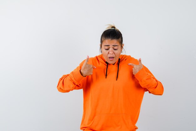 kobieta wskazująca na siebie w pomarańczowej bluzie z kapturem i wyglądająca na zaniepokojoną