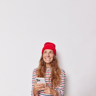 Kobieta wygląda marzycielsko powyżej trzyma telefon komórkowy używa aplikacji komórkowej do czatowania online z przyjaciółmi nosi czerwony kapelusz i swobodny sweter na białej ścianie studia