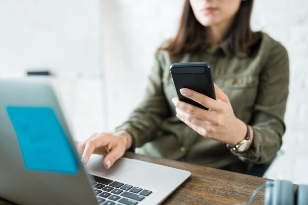 Kobieta wykonująca wielozadaniowość z telefonem komórkowym i laptopem przy biurku w biurze