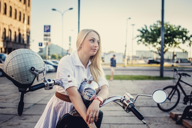 Bezpłatne zdjęcie kobieta z długimi blond włosami w białej sukni pozowanie w pobliżu skuter na ulicy w mieście.