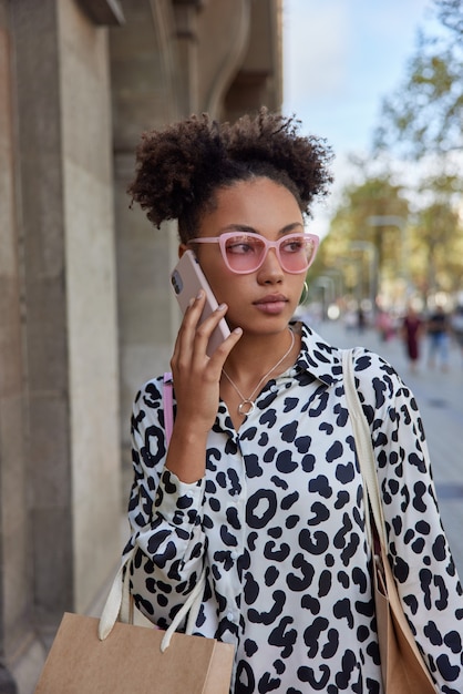 kobieta z dwoma bułeczkami ma na sobie modne różowe okulary przeciwsłoneczne i modną koszulę nosi papierową torbę po zakupach rozmawia telefonicznie przez smartfona pozuje na zewnątrz