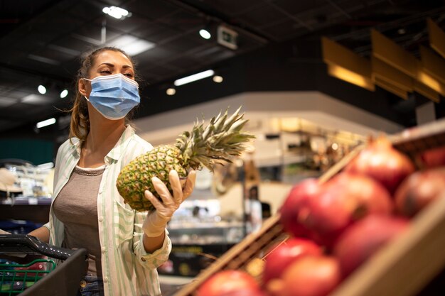 Kobieta z higieniczną maską i gumowymi rękawiczkami oraz koszyk w sklepie spożywczym, kupujący owoce podczas koronawirusa i przygotowujący się do kwarantanny pandemicznej