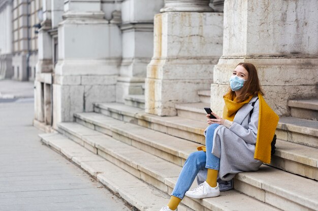 Kobieta z maską medyczną siedzi na schodach i za pomocą smartfona