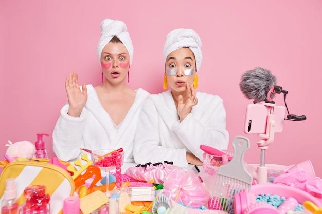 Bezpłatne zdjęcie kobiety noszą białe szlafroki i ręczniki na głowie zrecenzuj nagraj film o pielęgnacji skóry nałóż plastry upiększające pozuj przy stole z kosmetykami dookoła.
