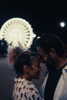 Kochająca para przytula się namiętnie na ulicy przed oświetlonym diabelskim młynem w nocy. para romansuje i spędza czas na świeżym powietrzu podczas wakacji w nocy