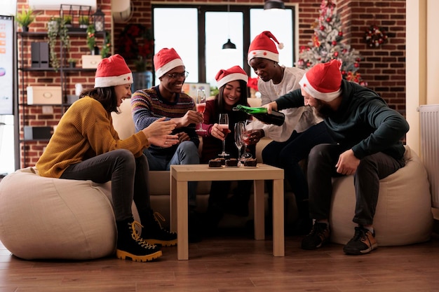 Koledzy nalewają kieliszki wina podczas świąt Bożego Narodzenia, czując się świątecznie i szczęśliwie na imprezie w wigilię Bożego Narodzenia w miejscu pracy z świątecznymi ozdobami. Picie alkoholu na zimowej imprezie.