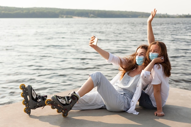 Bezpłatne zdjęcie koleżanki biorące selfie nad jeziorem z maseczkami medycznymi i rolkami