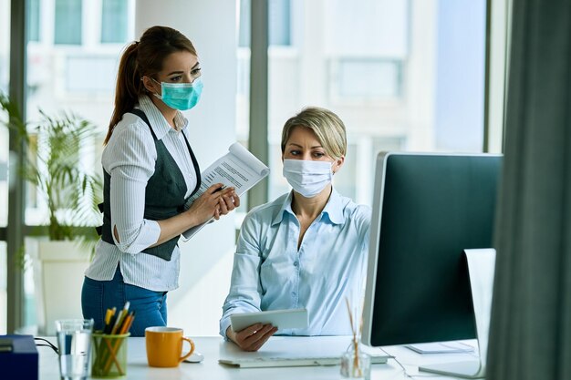 Koleżanki z pracy z maskami na twarz pracujące w biurze i czytające e-maile na komputerze