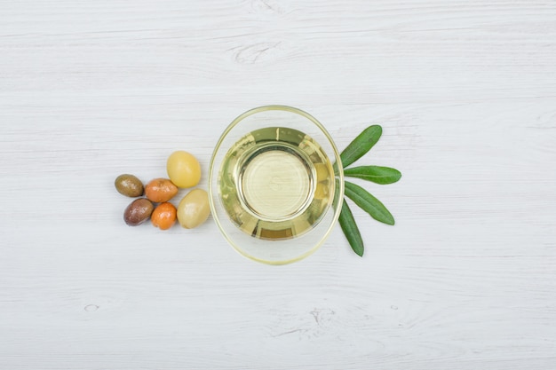 Kolorowe oliwki i oliwa z oliwek z oliwnymi liśćmi w szklanej puszce na białej drewnianej desce, odgórny widok.