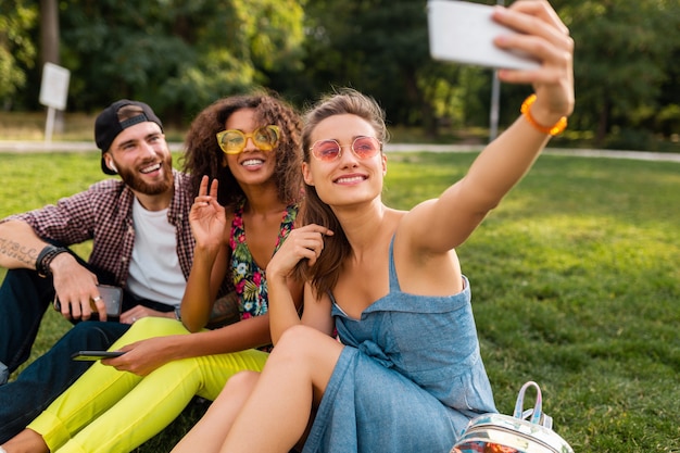 Kolorowe stylowe szczęśliwe młode towarzystwo przyjaciół siedzących w parku, mężczyzny i kobiet, którzy razem bawią się