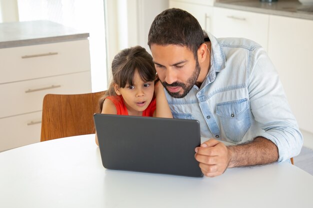 Koncentruje się podekscytowana dziewczyna i jej tata za pomocą laptopa, siedząc przy stole, patrząc na wyświetlacz.