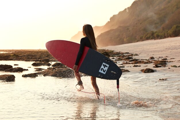 Koncepcja aktywnego sportu i stylu życia. Boardsurfer w ruchu, nosi czerwoną deskę surfingową, wbiega do wody, spędza wolny czas na surfowaniu