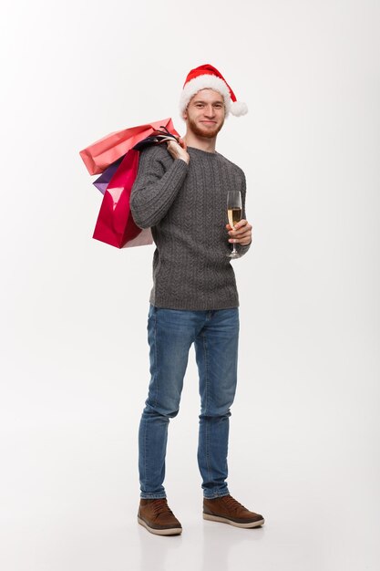 Koncepcja bożonarodzeniowa Młody przystojny mężczyzna z brodą trzymający kieliszek szampana i torby na zakupy ze szczęśliwym wyrazem twarzy