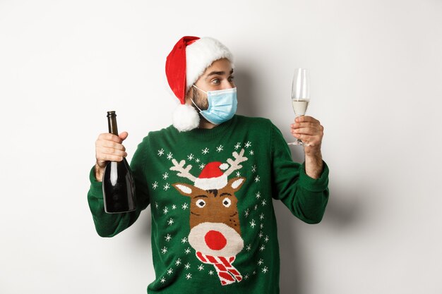 Koncepcja covid-19 i świąt Bożego Narodzenia. Mężczyzna w masce medycznej i kapeluszu Santa cieszący się lampką szampana, świętujący Nowy Rok, stojący na białym tle