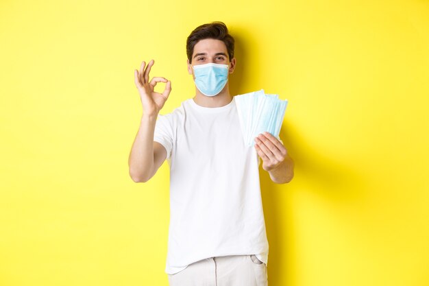 Koncepcja COVID-19, kwarantanny i środków zapobiegawczych. Zadowolony mężczyzna pokazujący dobry znak i dający maski medyczne, stojący na żółtym tle.