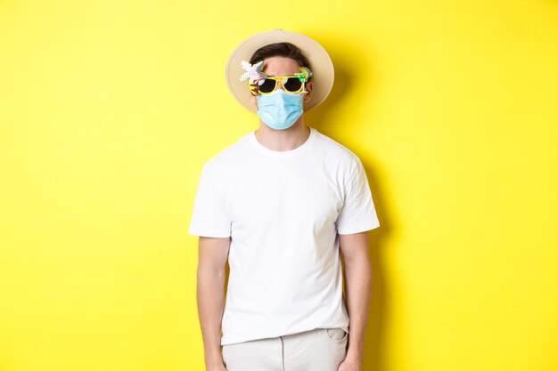 Koncepcja COVID-19, wakacje i dystans społeczny. Turysta mężczyzna w masce medycznej i letnim kapeluszu z okularami przeciwsłonecznymi, udaje się w podróż podczas pandemii, żółte tło.