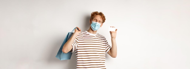 Bezpłatne zdjęcie koncepcja covid i stylu życia wesołego młodego człowieka z rudymi włosami nosi maskę medyczną pokazującą zakupy b