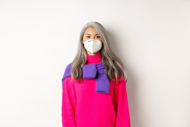 Koncepcja Covid, pandemii i dystansu społecznego. Stylowa azjatycka starsza kobieta nosząca respirator i patrząca na kamerę poważnie, stojąca na białym tle
