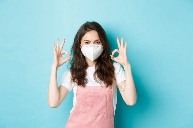 Koncepcja Covid, zdrowia i pandemii. Zadowolona piękna dziewczyna w respiratorze, medyczna maska pokazująca dobry znak zatwierdzenia, uisng środki zapobiegające koronawirusowi, niebieskie tło.