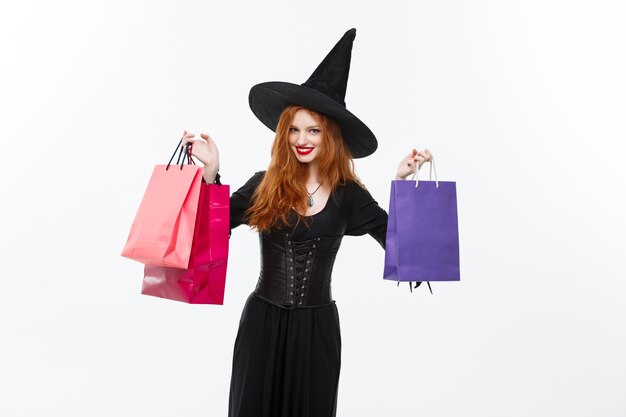Koncepcja czarownicy halloween szczęśliwa wiedźma halloween uśmiecha się i trzyma kolorowe torby na zakupy na białej ścianie