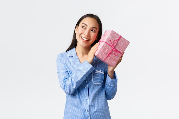 Koncepcja domu, wakacje i styl życia. Zaintrygowana dziewczyna z okazji urodzin w niebieskiej piżamie potrząsa pudełkiem z prezentem, aby dowiedzieć się, co jest w środku, zgadując prezent i uśmiechając się zaciekawiona, białe tło