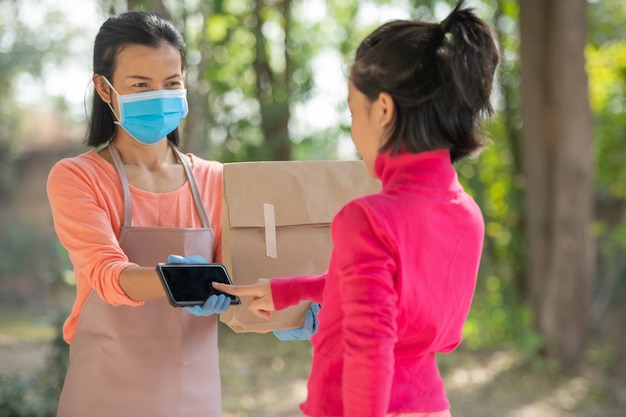 Koncepcja dostawy, poczty, ludzi i wysyłki. Młoda kobieta zaloguj się cyfrowy telefon komórkowy po otrzymaniu przesyłki od kuriera w domu. od dostawcy podczas epidemii COVID-19.