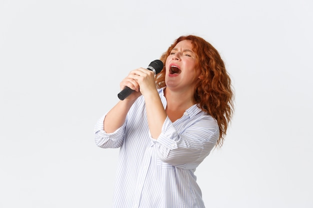 Koncepcja emocji, stylu życia i wypoczynku. Namiętna i beztroska rudowłosa performerka, kobieta w średnim wieku śpiewająca piosenkę do mikrofonu, piosenkarka grająca w karaoke, białe tło.