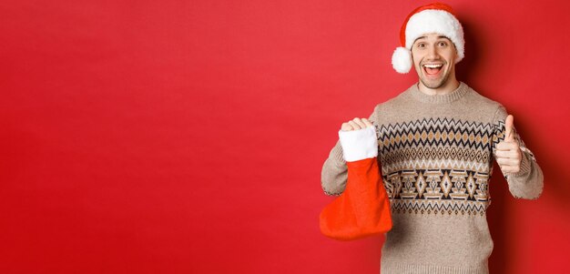 Koncepcja ferii zimowych nowego roku i uroczystości Wesoły przystojny mężczyzna w santa hat i sweter pokazano skarpetę świąteczną z cukierkami i prezentami, co thumbsup