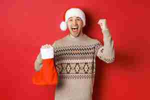 Bezpłatne zdjęcie koncepcja ferii zimowych, nowego roku i uroczystości. zdumiony i szczęśliwy mężczyzna krzyczący z radości, znalazł prezent w świątecznej pończosze i doping, podnosząc rękę do góry i uśmiechając się