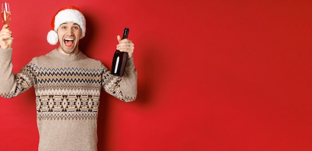 Koncepcja ferii zimowych, świąt bożego narodzenia i uroczystości. podekscytowany przystojny facet w swetrze i santa hat, ciesząc się przyjęciem sylwestrowym, podnosząc szklankę i butelkę szampana, bawiąc się