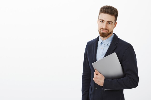 Koncepcja finansów, biznesu i technologii. Uroczy, elegancki młodzieniec z brodą i niebieskimi oczami w stylowym garniturze, trzymając laptop w ramieniu, uśmiechając się z pewnym siebie wyrazem twarzy