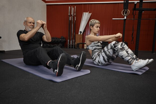 Koncepcja fitness, pracy zespołowej, sportu i szkolenia. Dwóch aktywnych sportowców starszy mężczyzna i młoda blondynka siedzą na matach i wykonują podkurczenia lub brzuszki podczas intensywnego treningu crossfit na siłowni
