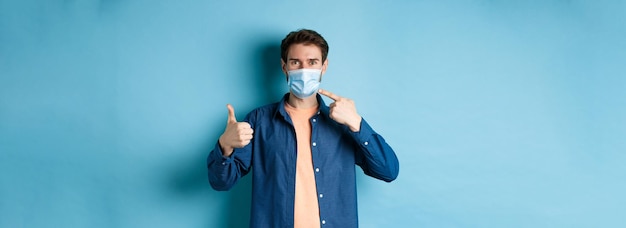 Bezpłatne zdjęcie koncepcja koronawirusa i pandemii młody zdrowy mężczyzna wskazujący na maskę medyczną i pokazujący nam kciuki