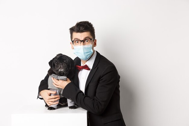 Koncepcja koronawirusa, zwierząt domowych i uroczystości. Szczęśliwy właściciel psa w garniturze i masce na twarz, przytulający słodkiego czarnego mopsa w kostiumie, stojący na białym tle