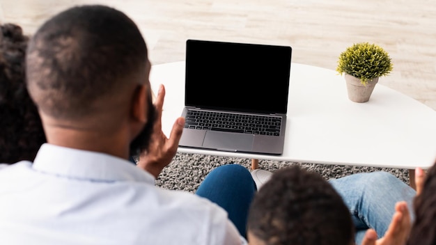 Koncepcja ludzi i technologii. przez ramię widok młodej afroamerykańskiej rodziny korzystającej z laptopa z pustym pustym ekranem dla szablonu makiety, siedzącej na kanapie w salonie w domu