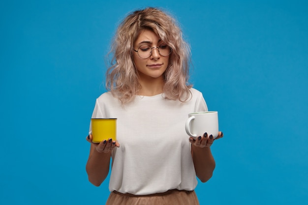 Bezpłatne zdjęcie koncepcja ludzi, stylu życia, napojów i żywności. zabawna niezdecydowana, wątpliwa młoda kobieta z rozczochranymi różowawymi włosami stoi przed dylematem, wahająca się przy wyborze między kawą a herbatą