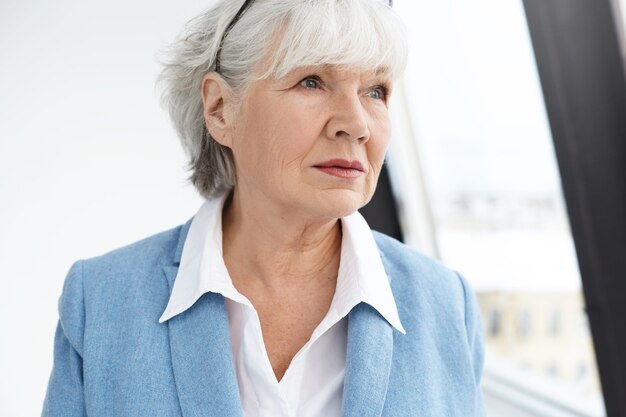 Koncepcja ludzi, wieku, stylu życia, mody i emerytury. Zdjęcie eleganckiej modnej sześćdziesięcioletniej bizneswoman z pomarszczoną twarzą i siwymi włosami, zastanawiając się nad sprawami biznesowymi, pozując przy oknie